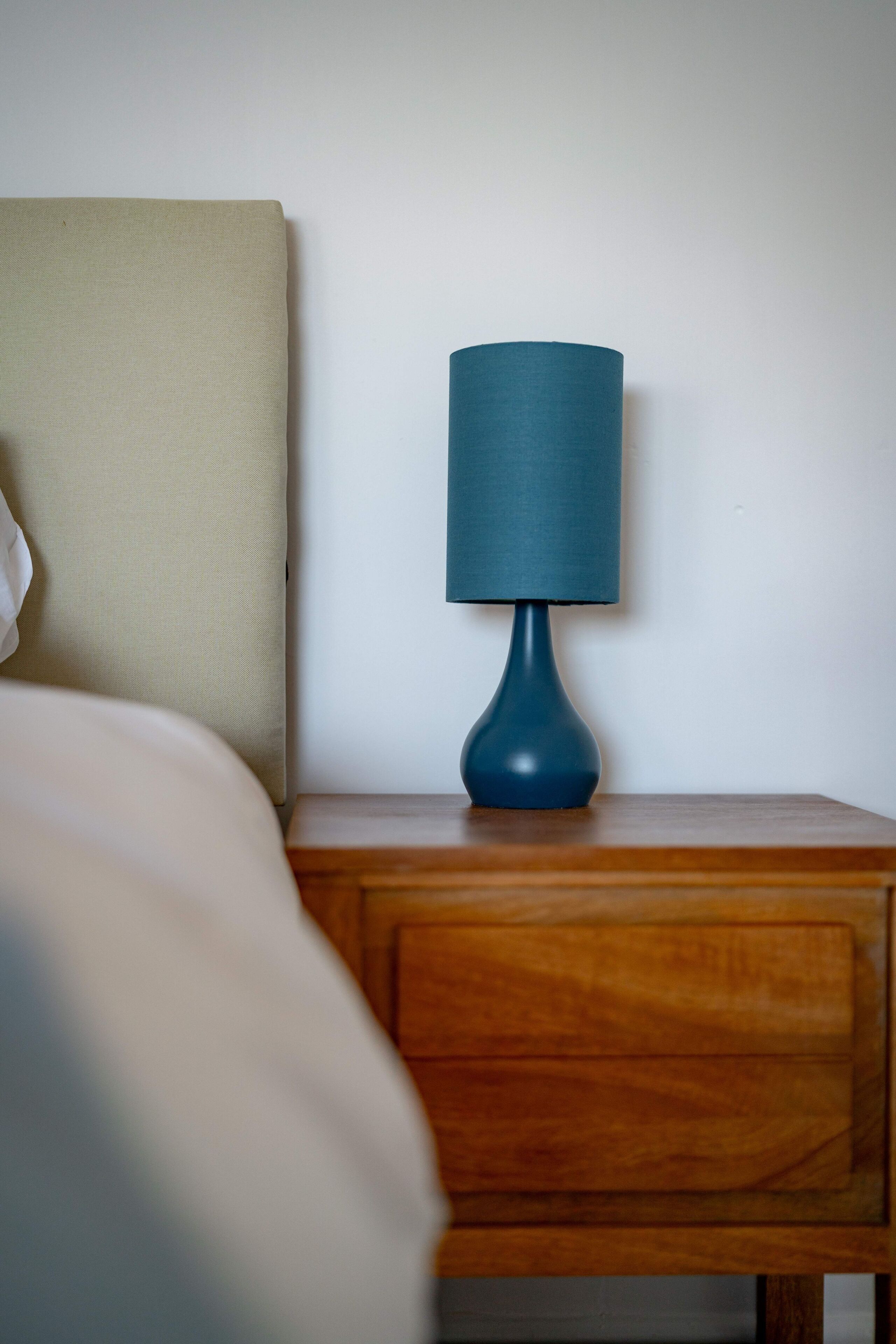 Ein Nachttisch aus Holz mit einer blauen Lampe und einem beigen Kissen vor einer schlichten Wand.
