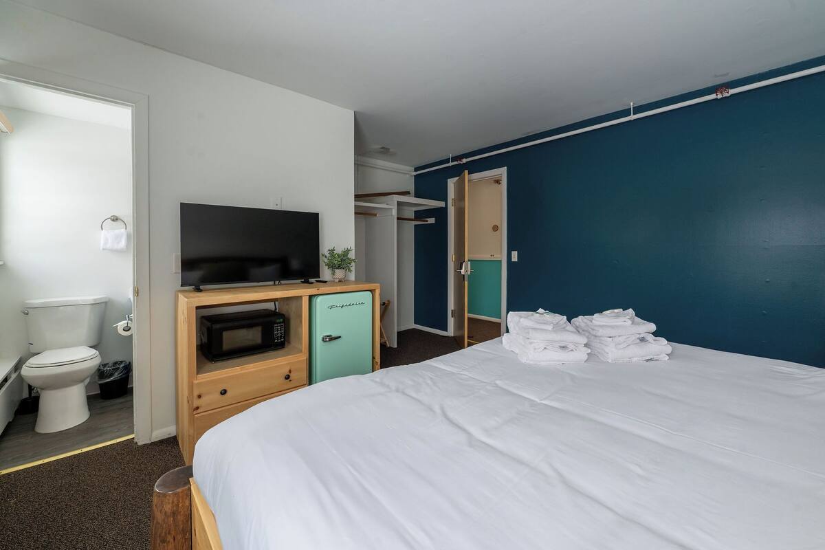 A hotel room with a bed, a microwave, a television, a small cabinet, a toilet, and a blue wall.