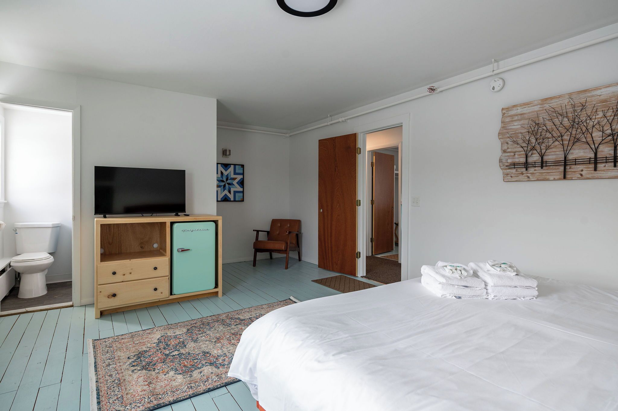 A bedroom with a bed, a television on a wooden cabinet, a chair, and a painting on the wall.