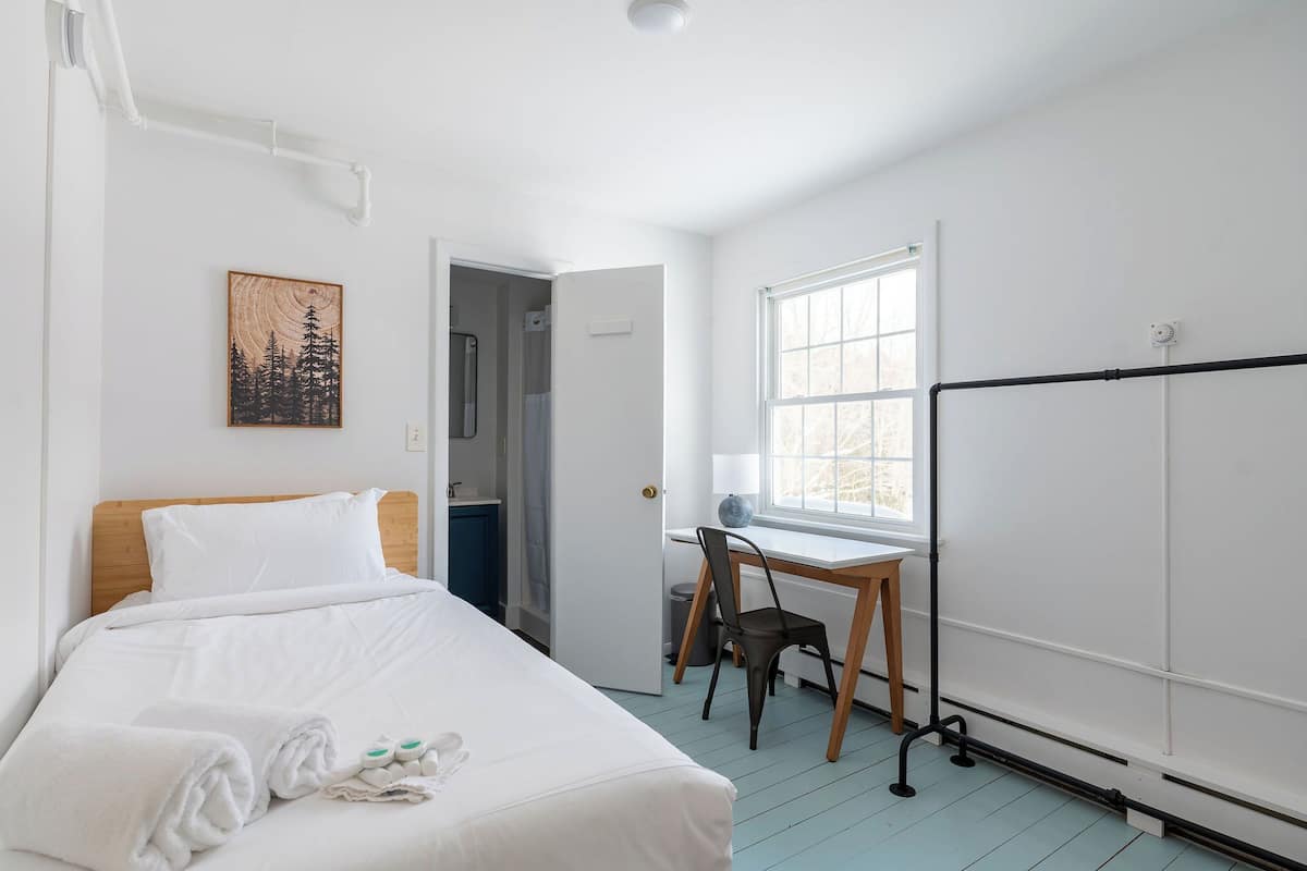 A hotel room with a bed, a desk, a chair, a window, and a painting on the wall.