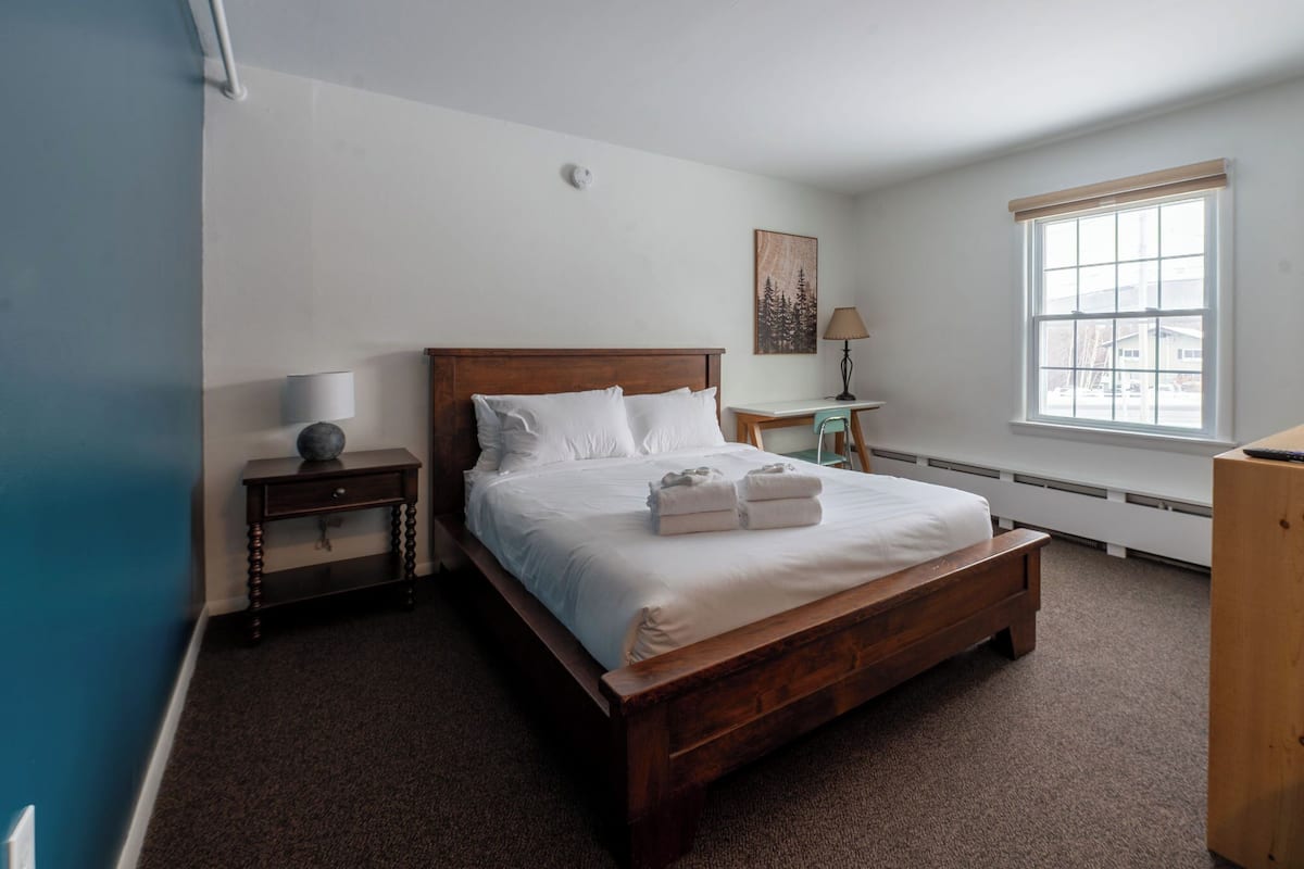 A bedroom with a wooden bed, a nightstand, a desk, a lamp, and a window.