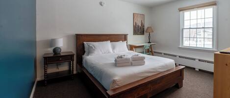 Une chambre à coucher avec un lit en bois, une table de chevet, un bureau, une lampe et une fenêtre.