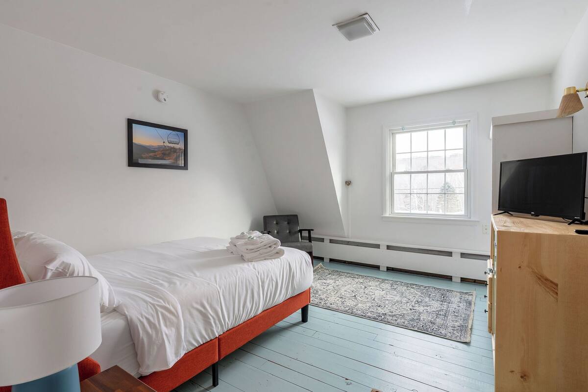 A bedroom with a bed, a TV, a window, and a painting on the wall.