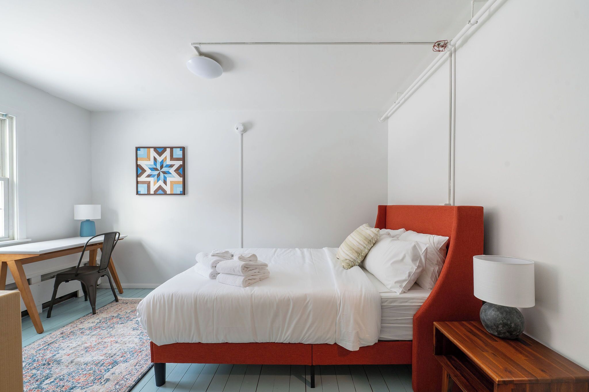 A neatly arranged bedroom with a bed, bedside table, lamp, and a desk with chair.