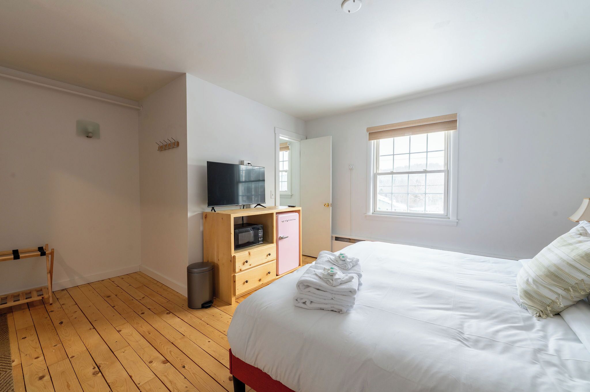 A bedroom with a wooden floor, a bed with white bedding, a wooden wardrobe, a television on top, and a window with blinds.
