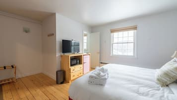 A bedroom with a wooden floor, a bed with white bedding, a wooden wardrobe, a television on top, and a window with blinds.