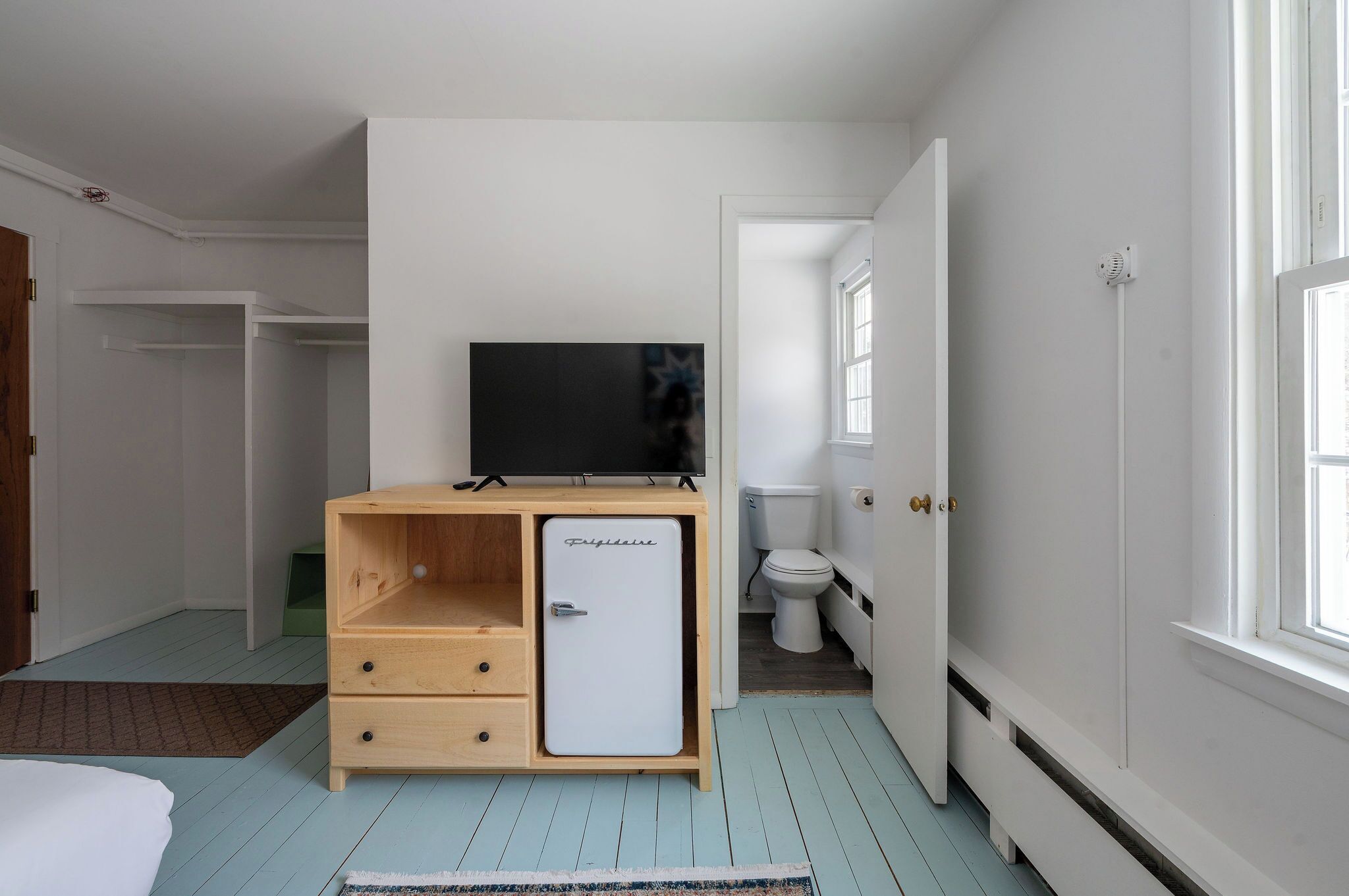 Quarto compacto com cama, um pequeno armário de madeira com televisão, um vaso sanitário branco e uma janela que oferece luz natural.
