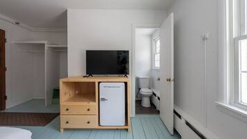 Una habitación pequeña con una cama, un mueble de madera con un televisor, un inodoro blanco y una ventana que deja entrar luz natural.