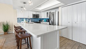 Uma cozinha moderna com armários brancos, uma ilha grande com banquetas, eletrodomésticos de aço inoxidável e iluminação pendente.