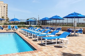 A poolside area with lounge chairs and umbrellas, a swimming pool, and a building in the background. - Gulf View, Gourmet Kitchen: Suntide III - Unit 203 (South Padre Island)