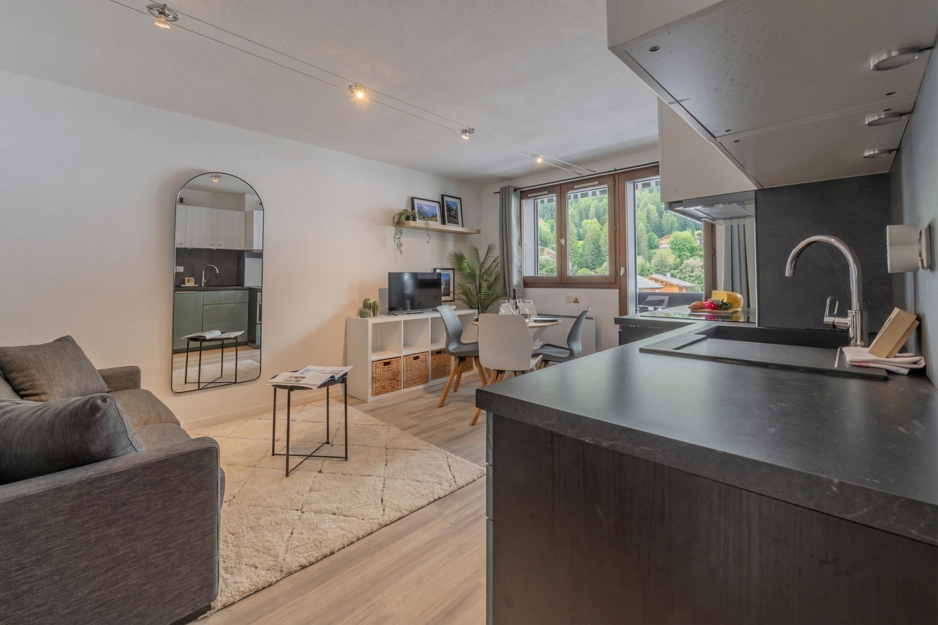 A modern kitchen with a dining area, a sofa, and a large window with a view of greenery.