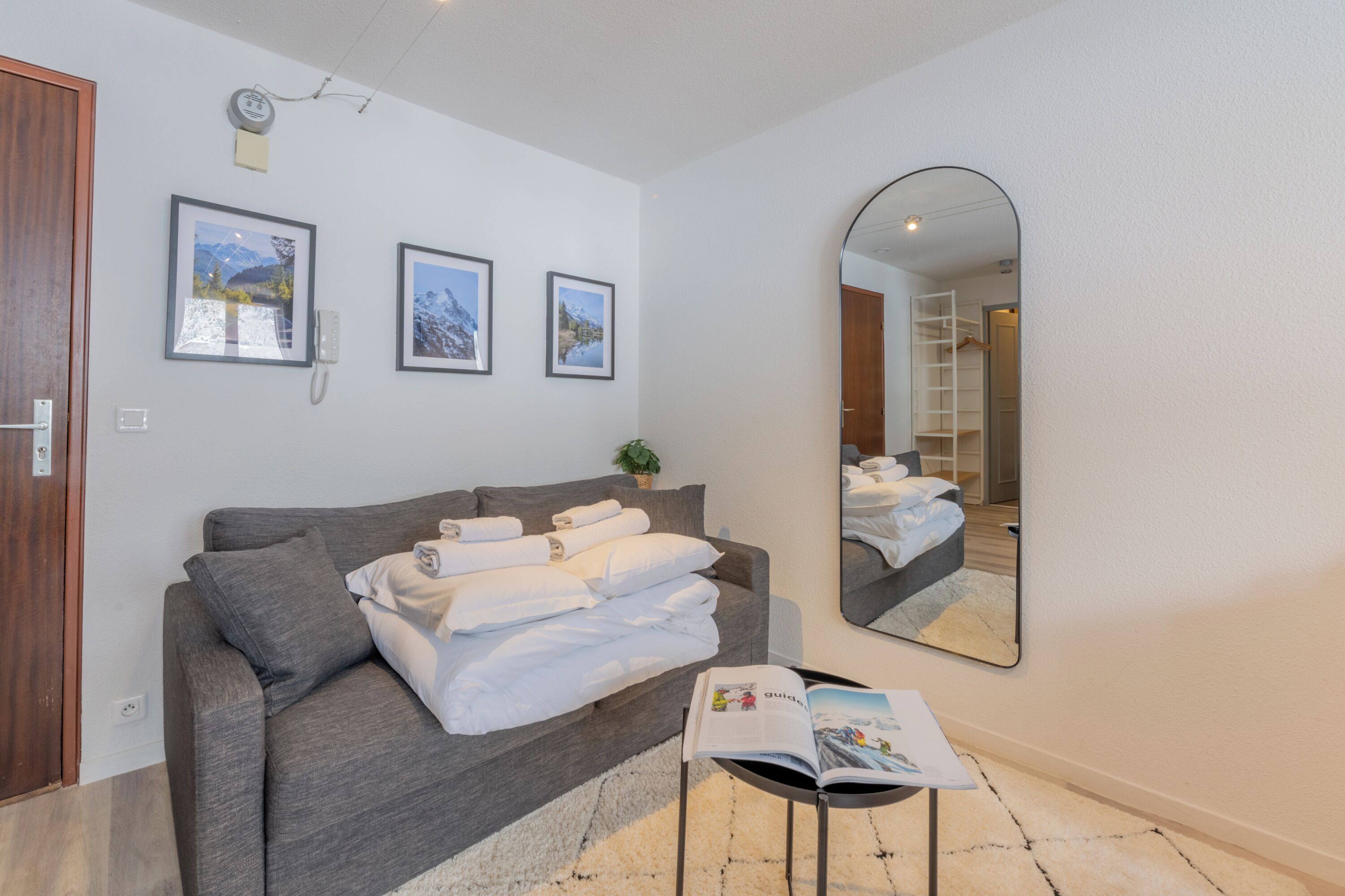 A modern hotel room with a grey sofa, a large mirror, and three framed pictures on the wall.