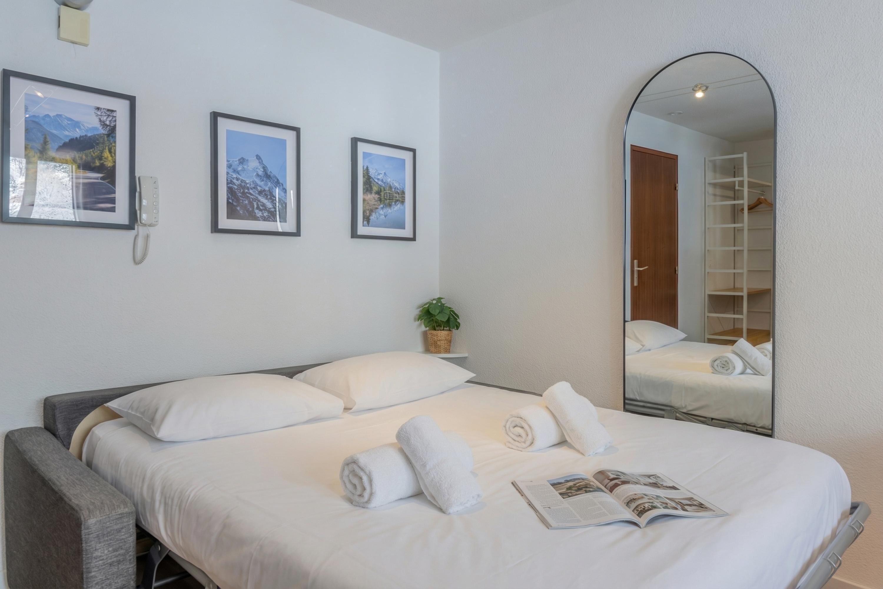 A neatly made bed with white linens and rolled towels, a bedside plant, and a mirror reflecting the room.