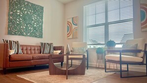 Un salon avec un canapé en cuir marron, une table basse en bois et un fauteuil beige. Les murs sont ornés de tableaux abstraits et une grande fenêtre est dotée de stores.