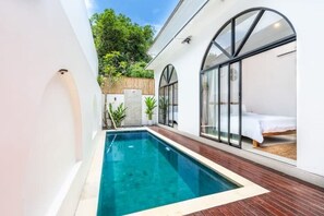 Un espace piscine moderne avec une piscine à eau claire, une terrasse en bois et un lit recouvert d’une courtepointe blanche.