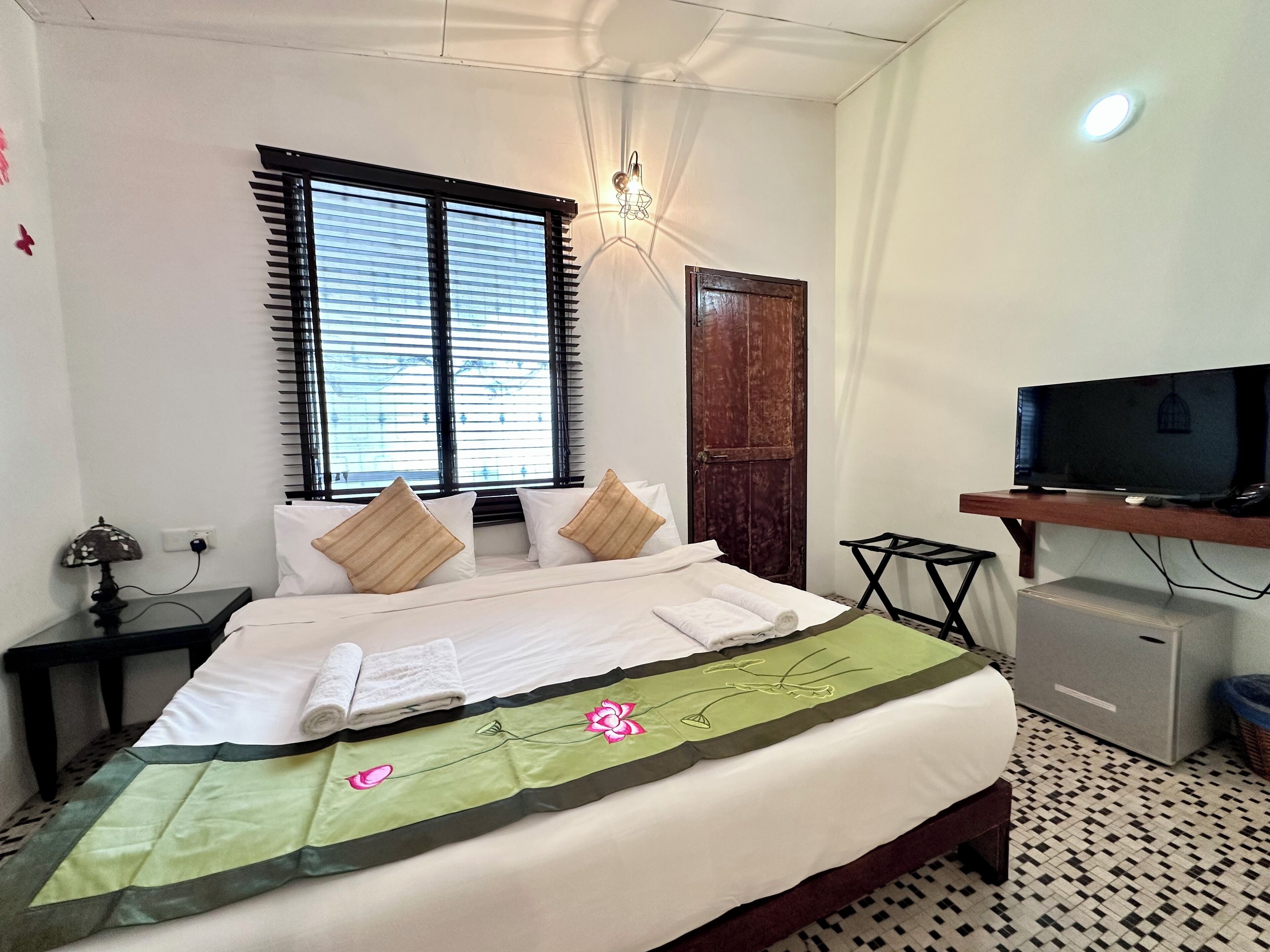 Quarto de hotel com cama, televisão em suporte, janela com persianas, porta e luminária no teto.