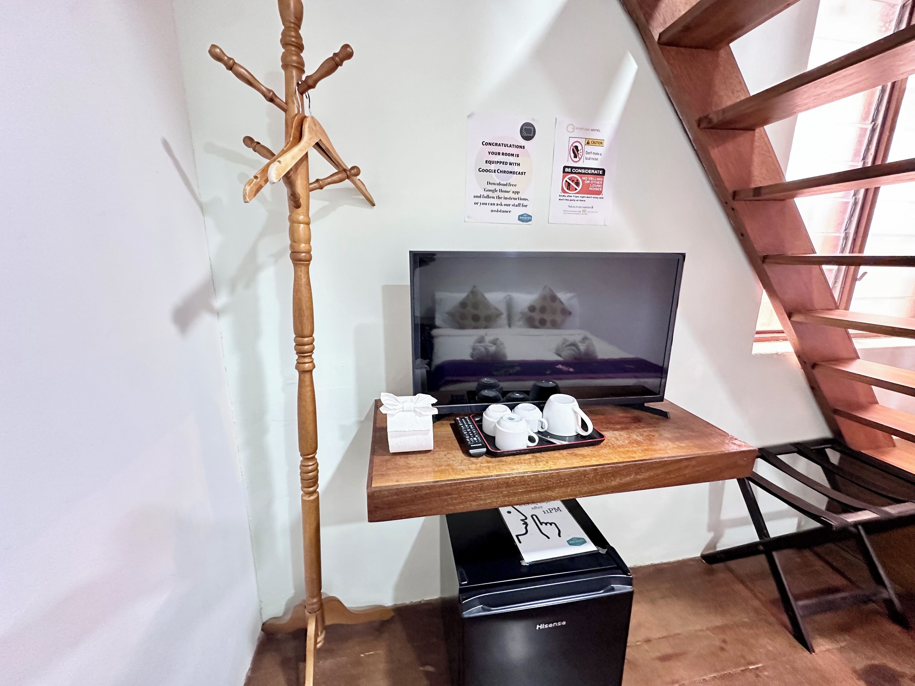 A hotel room with a wooden coat rack, a flat-screen TV, a tray with cups and a remote, and a black Hisense air purifier.