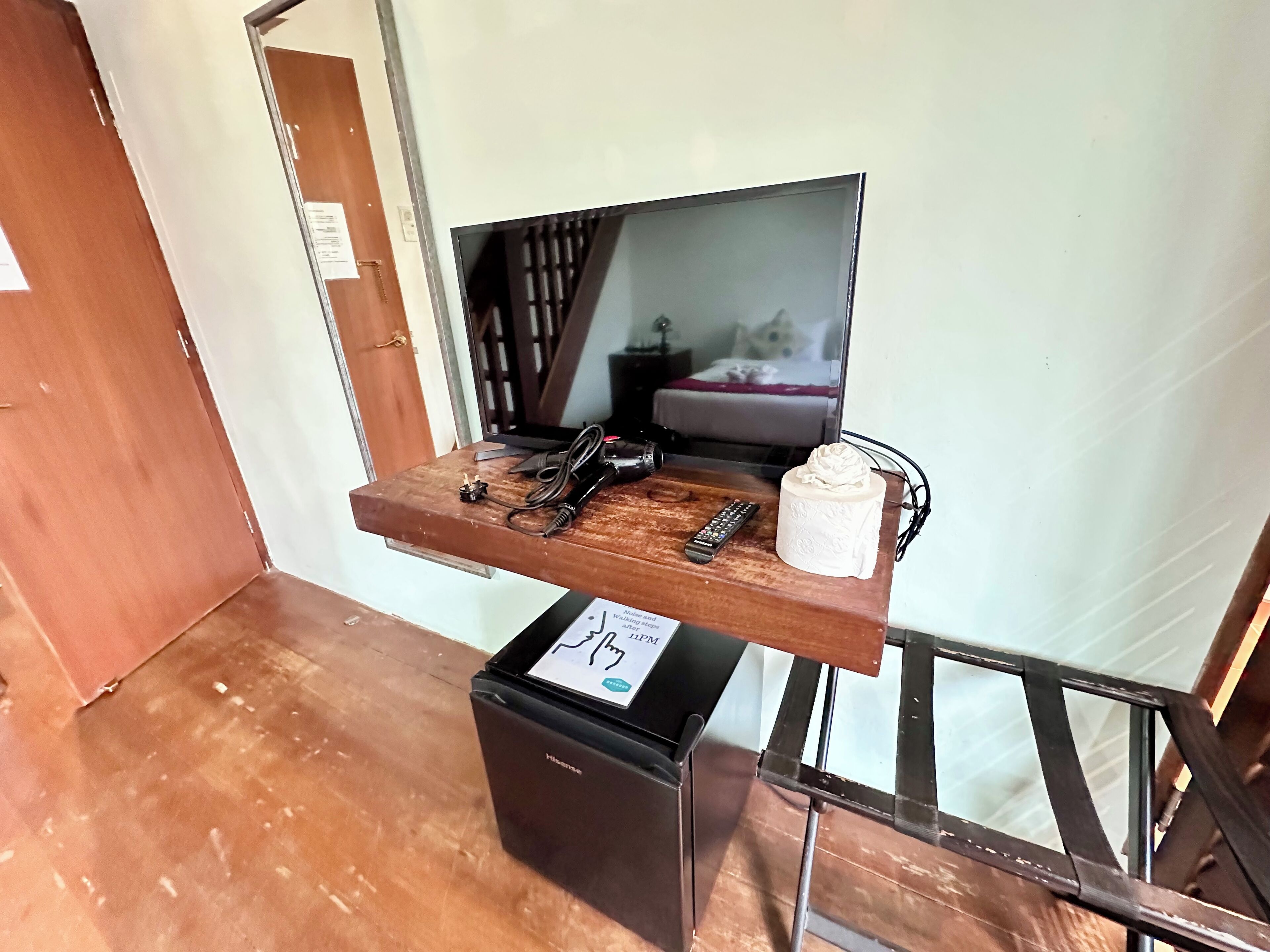 A compact room with a bed, a television on a wooden shelf, a small black appliance, and a remote control.