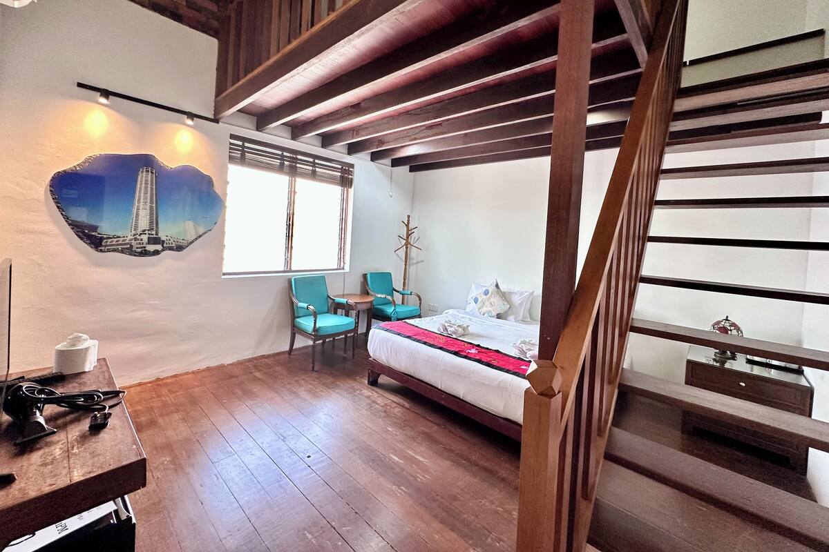 A room with a wooden staircase, a bed with a red blanket, two chairs, a coat rack, and a wall-mounted picture.