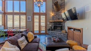 A spacious living room with a stone fireplace, a large TV mounted on the wall, and a cozy seating area with a sofa and ottomans. - Ski-in/ski-out 4-bedroom townhome in Crested Butte! (Gunnison)