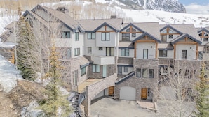 A multi-story residential building with a stone facade, multiple balconies, and a ski slope in the background. - Ski-in/ski-out 4-bedroom townhome in Crested Butte! (Gunnison)