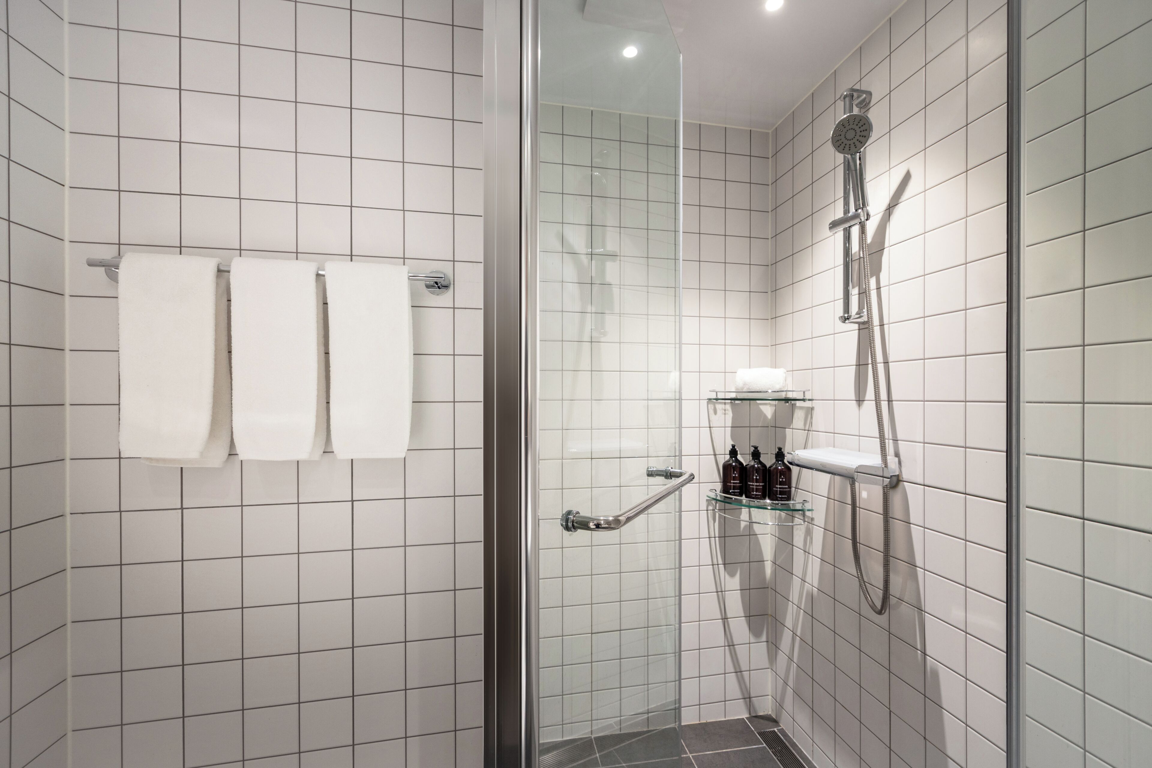 Une salle de bain moderne avec des carreaux blancs, une douche en verre, un porte-serviettes avec trois serviettes et une étagère contenant des articles de toilette.