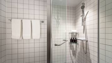 A modern bathroom with white tiles, a glass shower enclosure, a towel rack with three towels, and a shelf with toiletries.