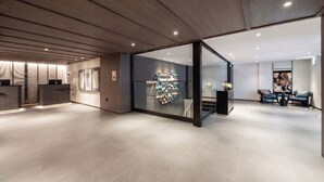 Un moderno vestíbulo de hotel con un mostrador de recepción, un área de descanso con sofá y mesa de centro, y un muro de vidrio con un arreglo decorativo.