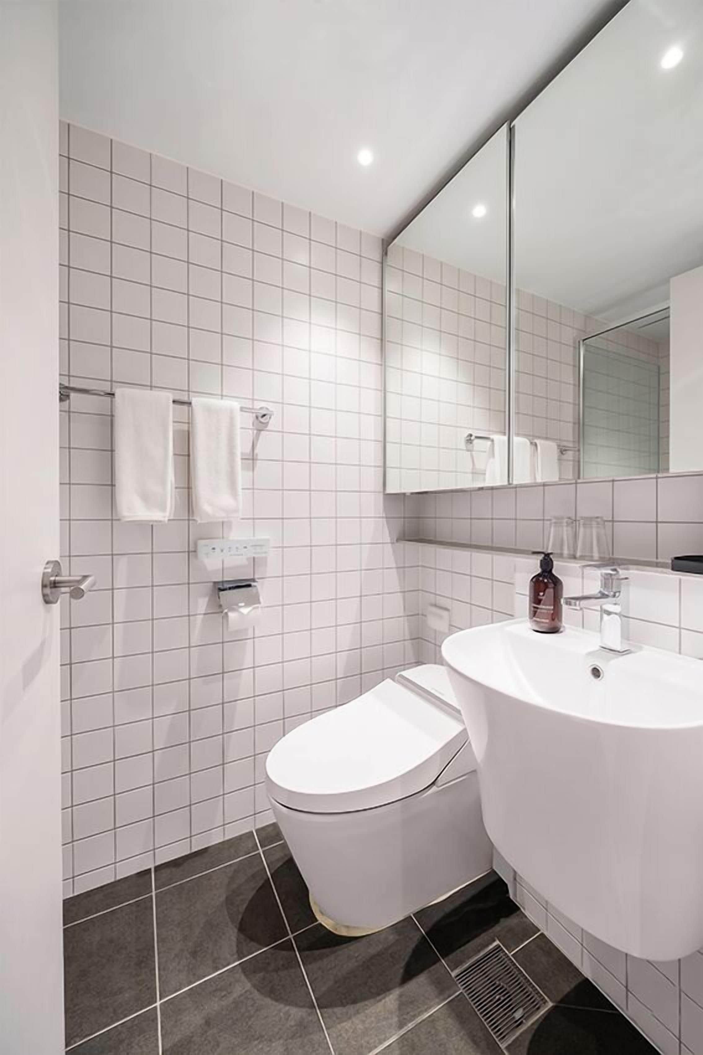 A modern bathroom with white tiles, a toilet, a sink, a mirror, and a towel rack.
