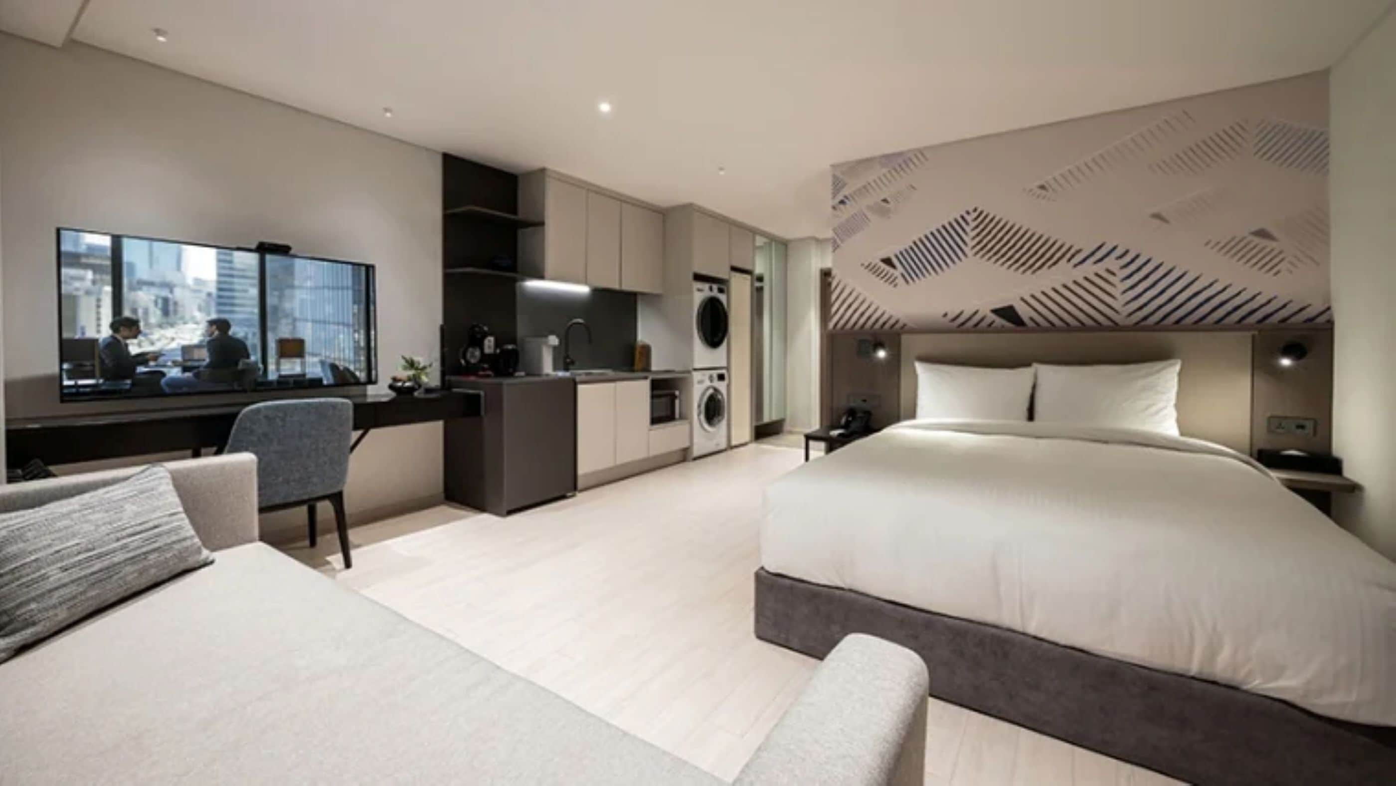 A modern hotel room with a large bed, a desk, a seating area, a kitchenette, and a view of the city.