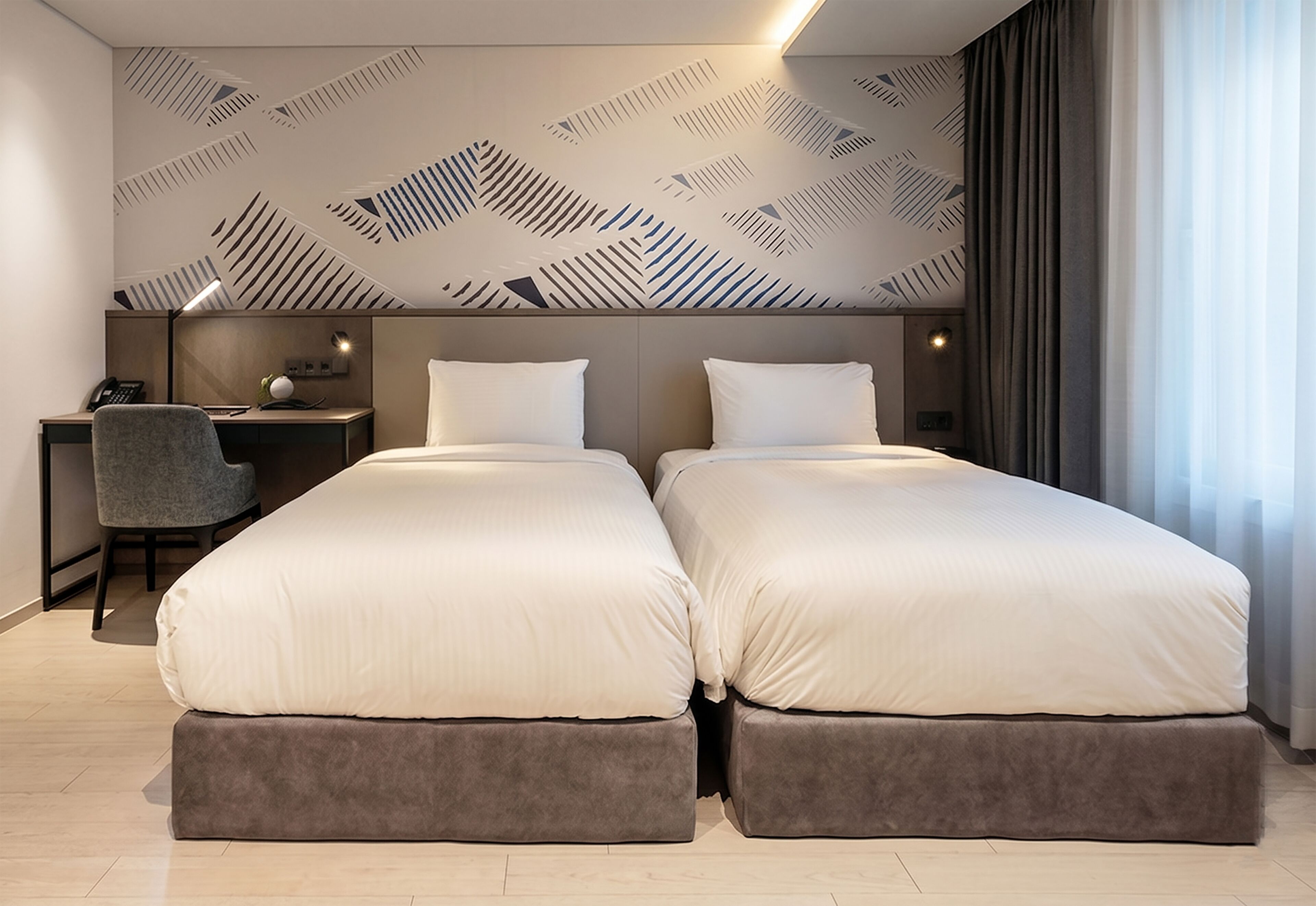 A hotel room with two beds, a desk with a chair, a patterned wall, and a window with curtains.