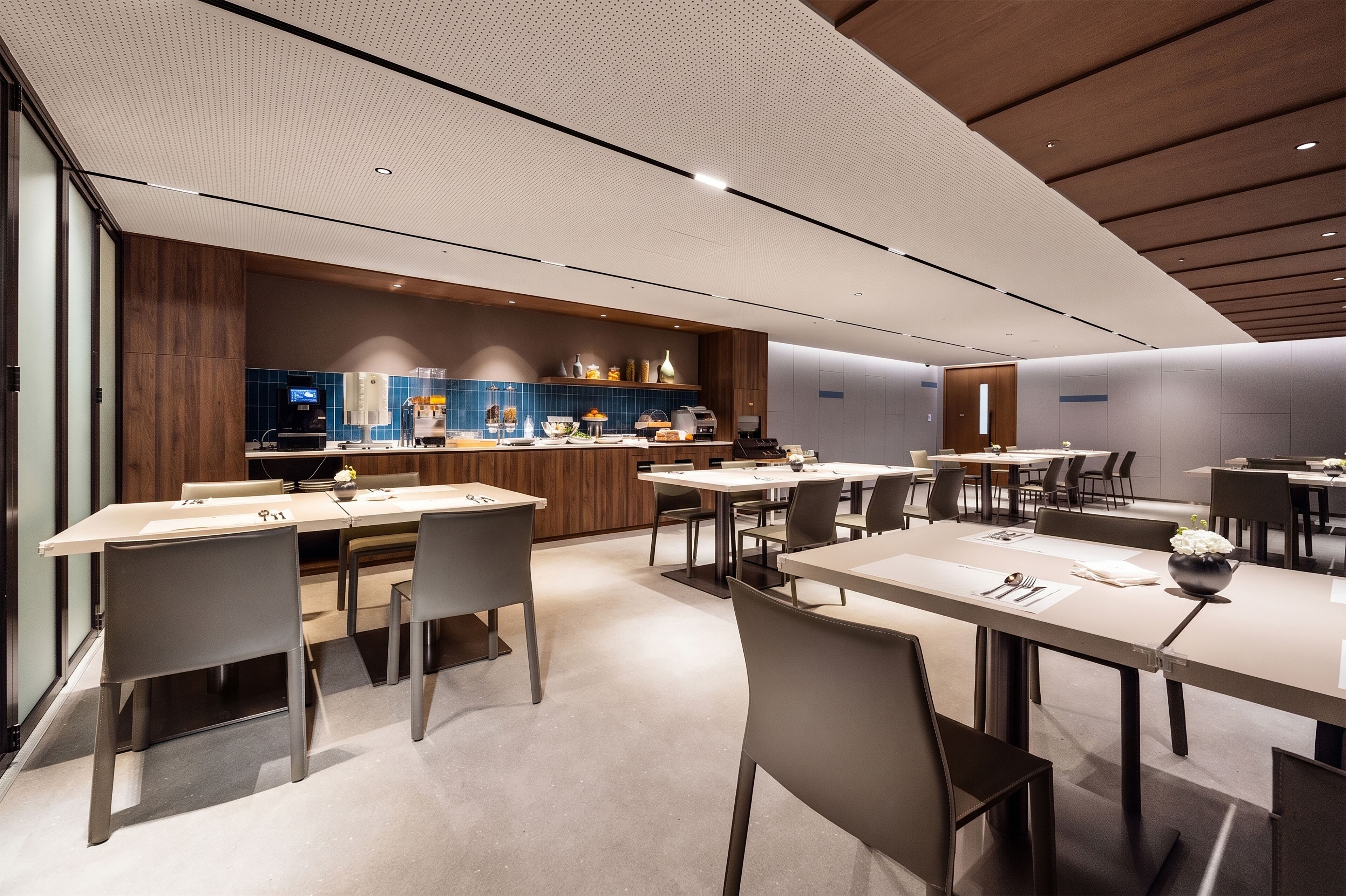 A modern hotel dining area with multiple tables and chairs, a kitchen area with blue-tiled backsplash, and wooden accents.