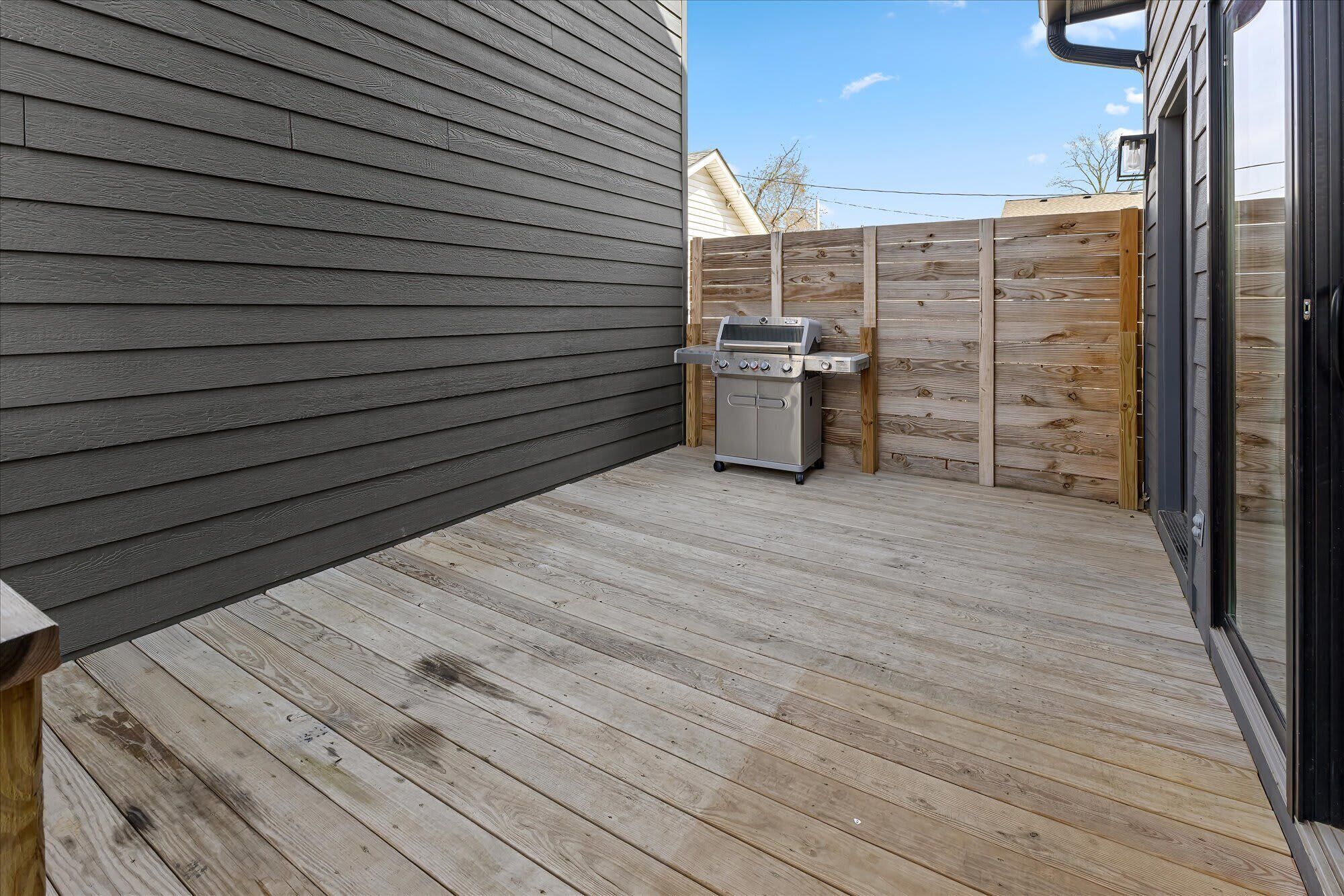 Una terraza de madera con una parrilla, una puerta corrediza de vidrio y un cerco de madera.