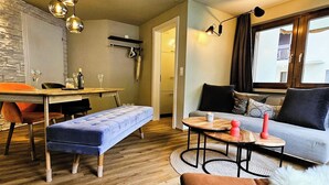 A modern living room with a sofa, a dining table, and a bench. There is a window with curtains and a view of buildings outside. - Achat 4 by Interhome (Saas-Fee)