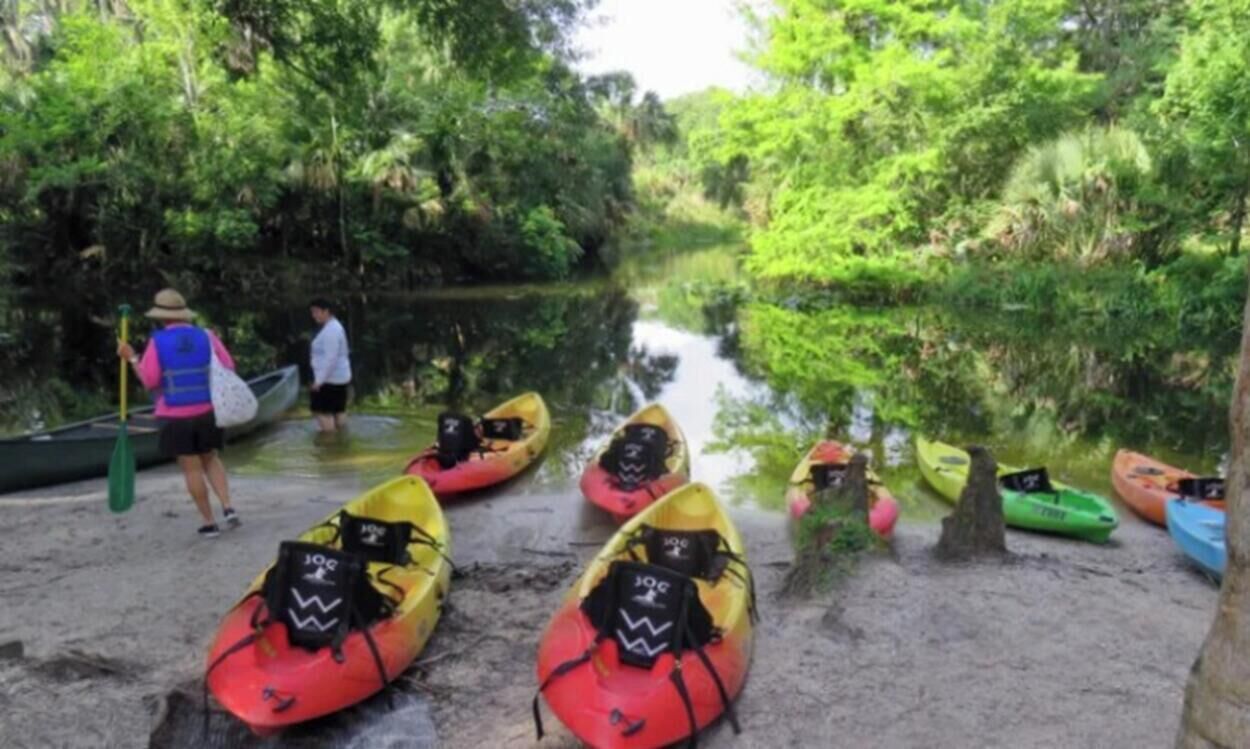 A river with kayaks lined up on the shore, a person in a life jacket, and a canoe in the background.