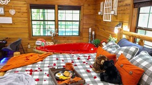 A cozy cabin room with a checkered floor, a red bathtub, a bed with a teddy bear, and a breakfast tray on the bed. - Heart Shaped Jacuzzi in main bedroom overlooking Fox Lake! Free Kayaks & Bikes (Angola)