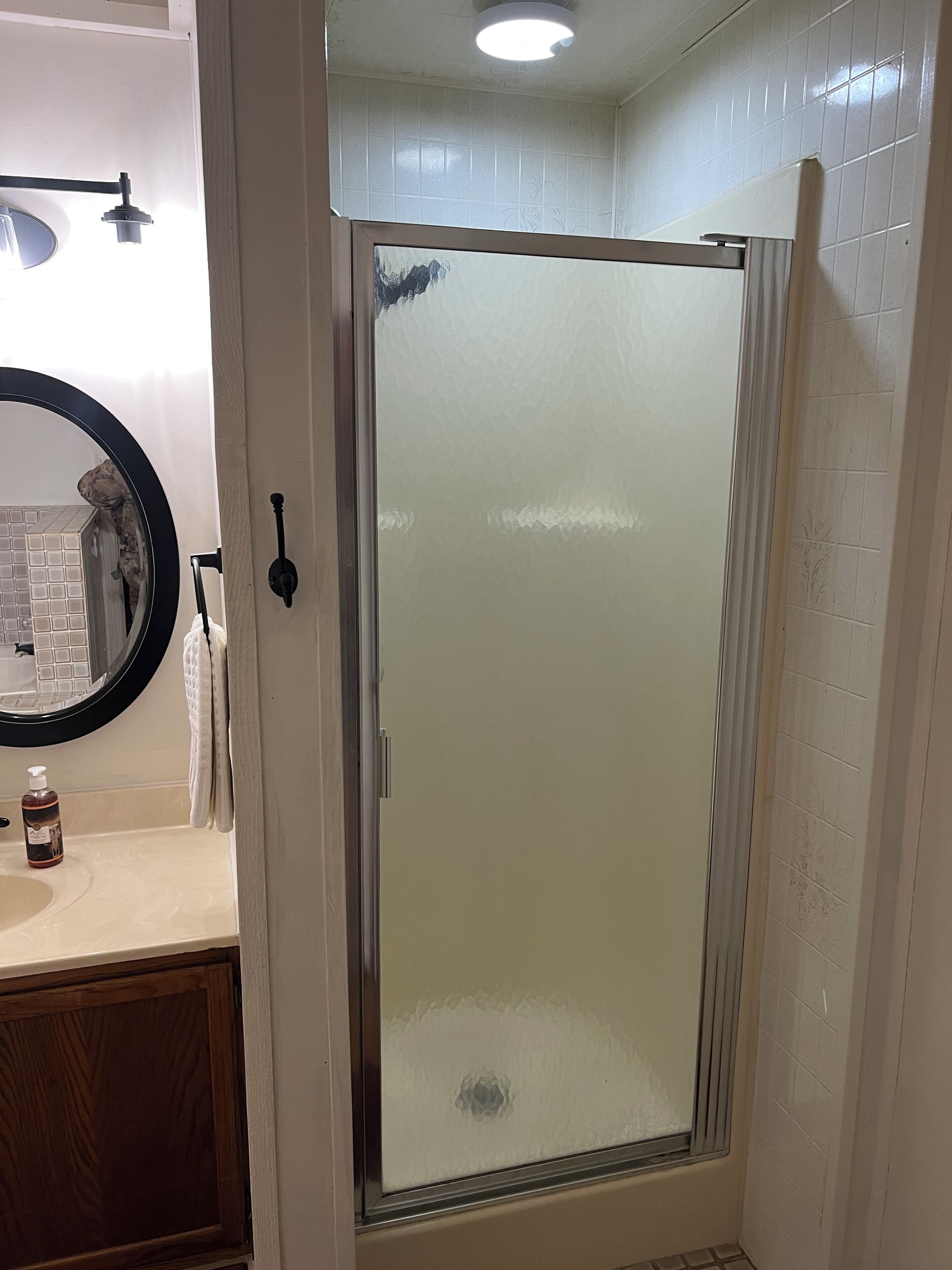 A bathroom with a glass-enclosed shower, a round mirror, a towel rack, and a bottle of hand soap on the countertop.
