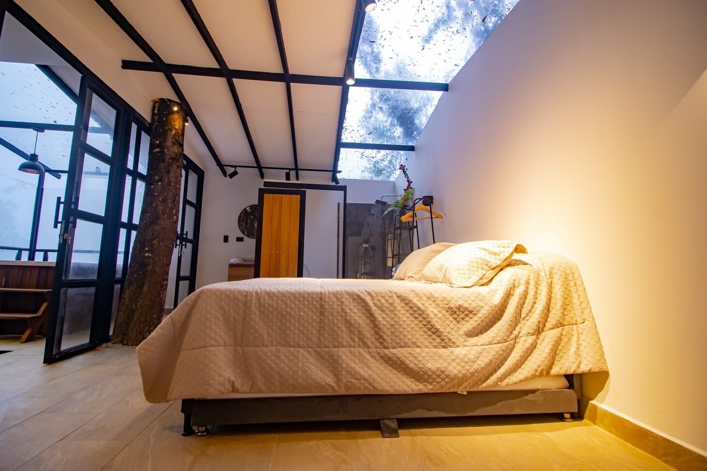 Un dormitorio con cama, un ventanal grande y el tronco de un árbol en la esquina.