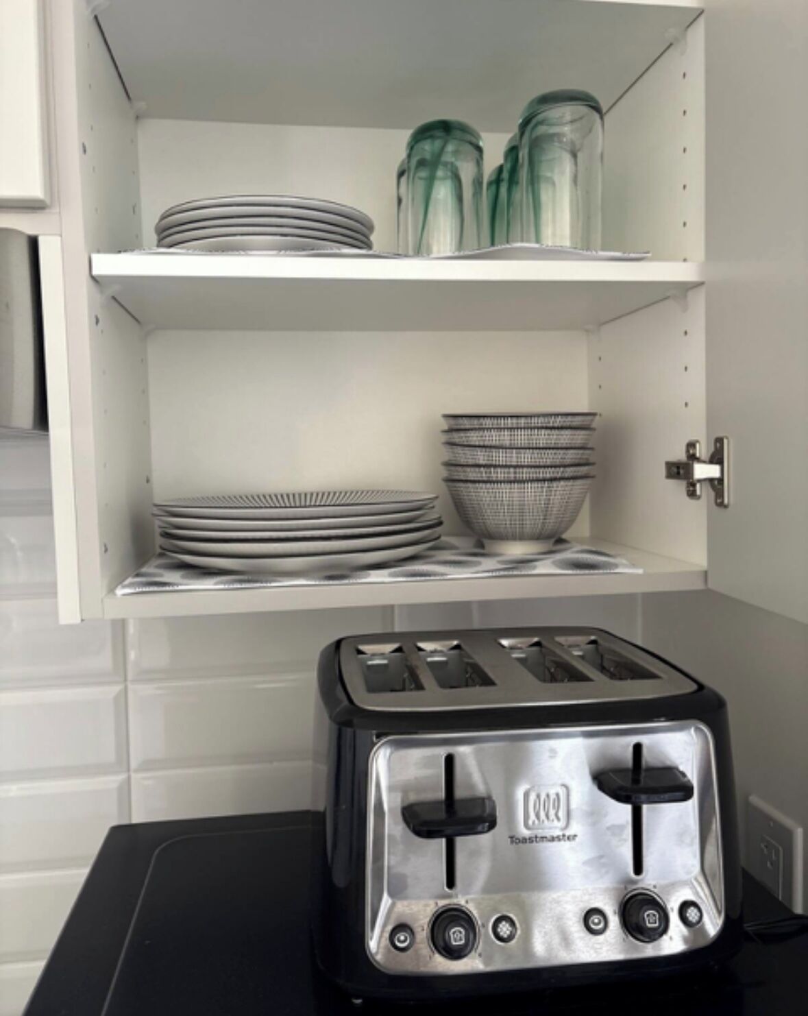 Eine Küche mit einem Toaster auf einer schwarzen Arbeitsplatte, weißen Regalen mit Tellern und Schüsseln und einer weißen, gefliesten Rückwand.