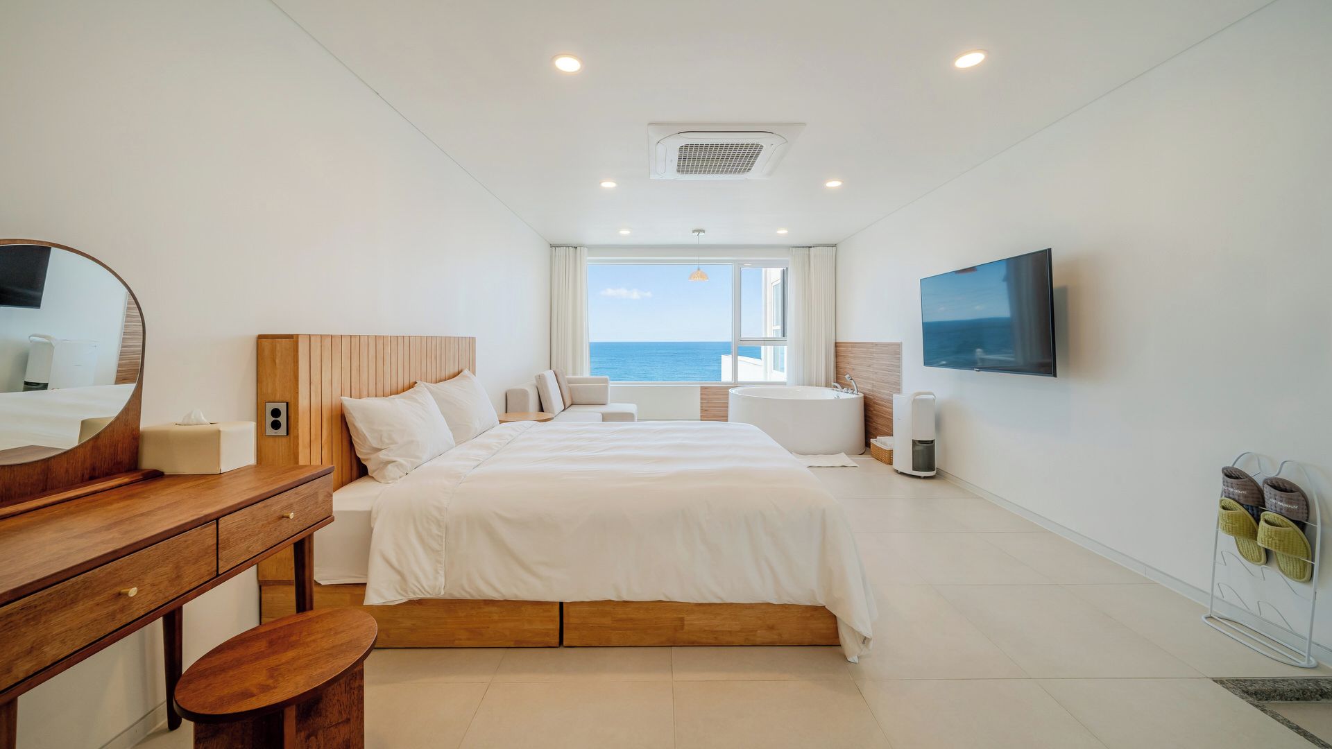 A hotel room with a large bed, a wooden nightstand, a desk, a chair, a TV, and a view of the sea.