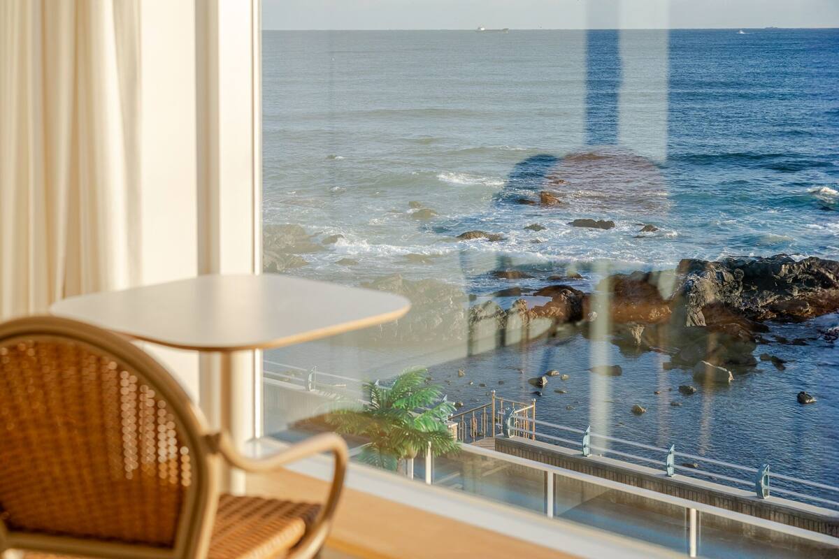 A room with a view of the ocean through a glass window, a chair, and a small table.