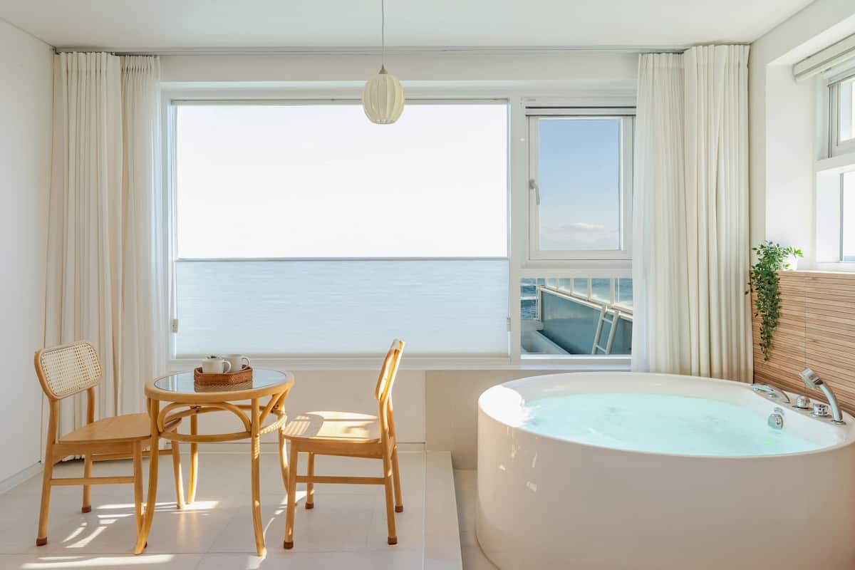 A modern bathroom with a large bathtub, a wooden chair, a small round table, and a view of the sea through a large window.