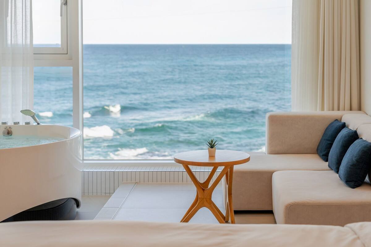 A room with a view of the ocean, a bathtub, a sofa with cushions, and a small round table.