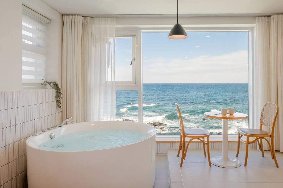A bathroom with a large bathtub, a small table with chairs, and a view of the ocean.