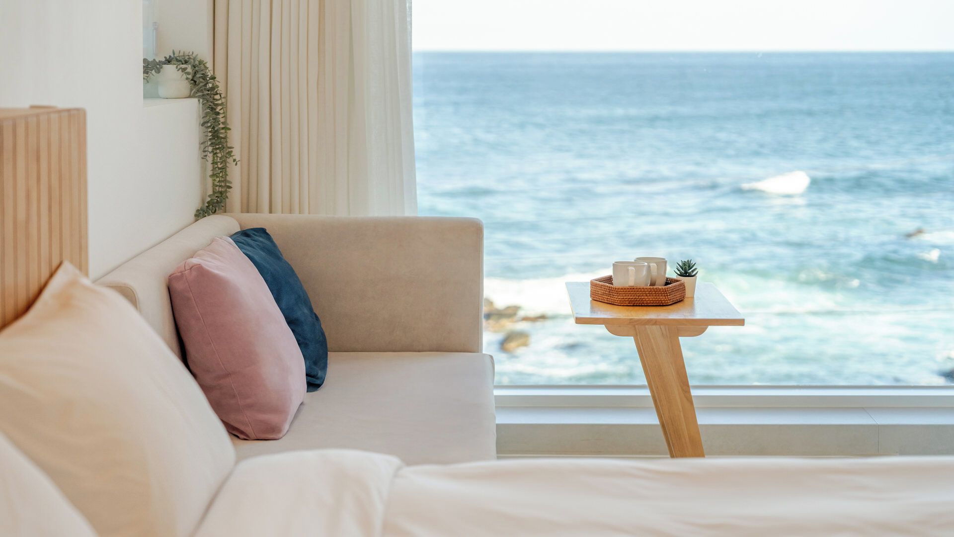 A bedroom with a view of the ocean, a wooden side table with a basket, and a beige sofa with pillows.