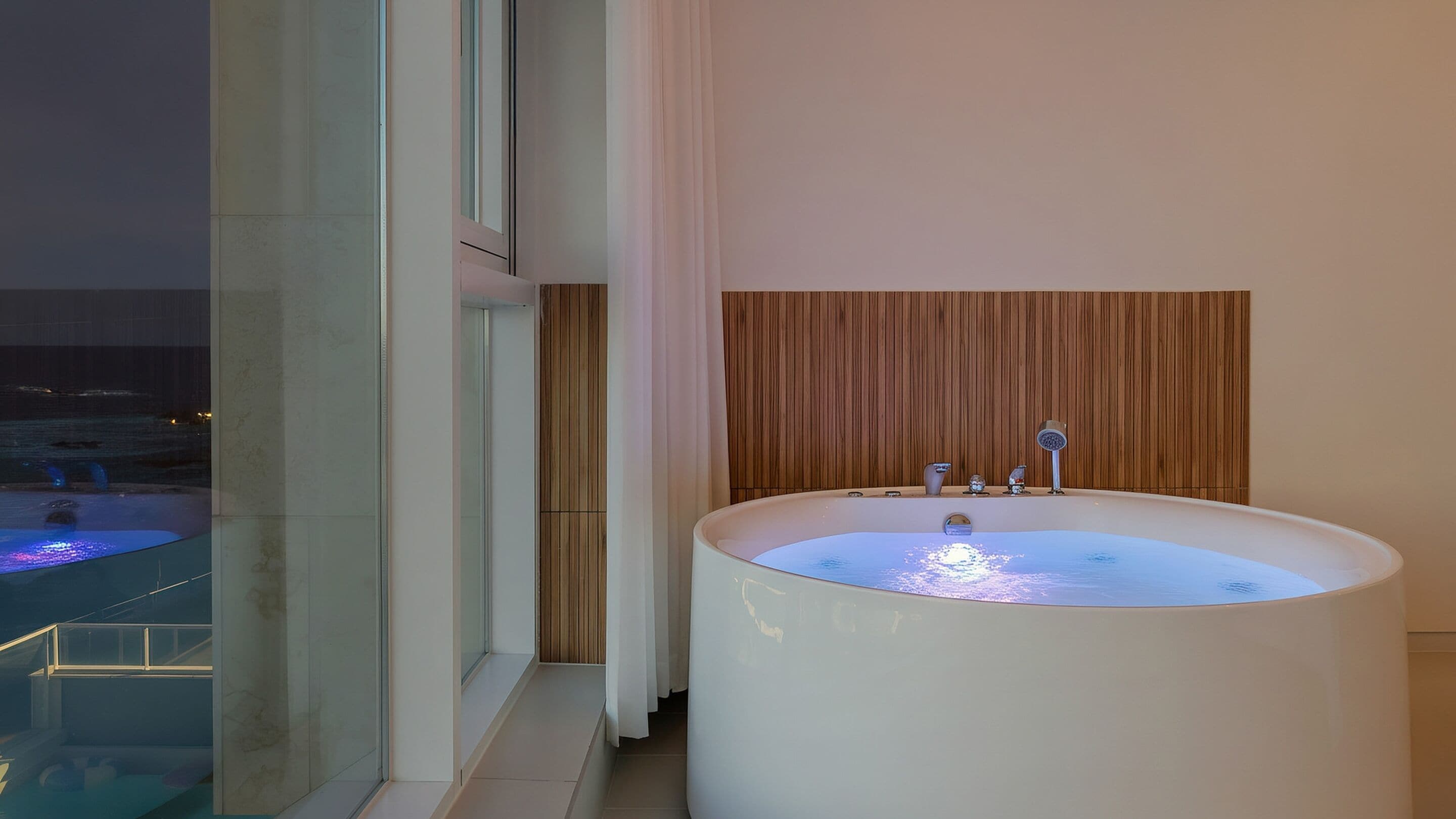 A modern bathroom with a large bathtub, a wooden panel wall, and a glass window overlooking a pool.