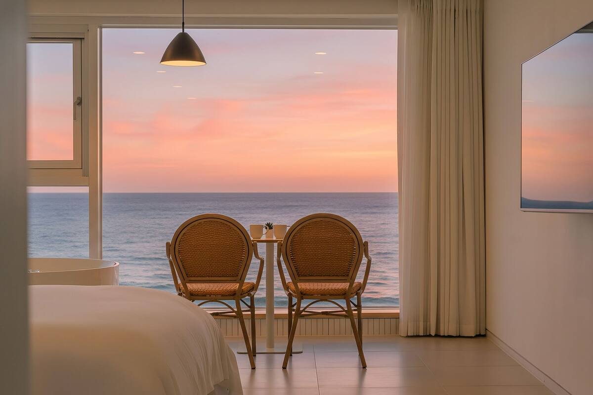 A room with a view of the ocean at sunset, featuring two wicker chairs, a bed, a TV, and a ceiling light.