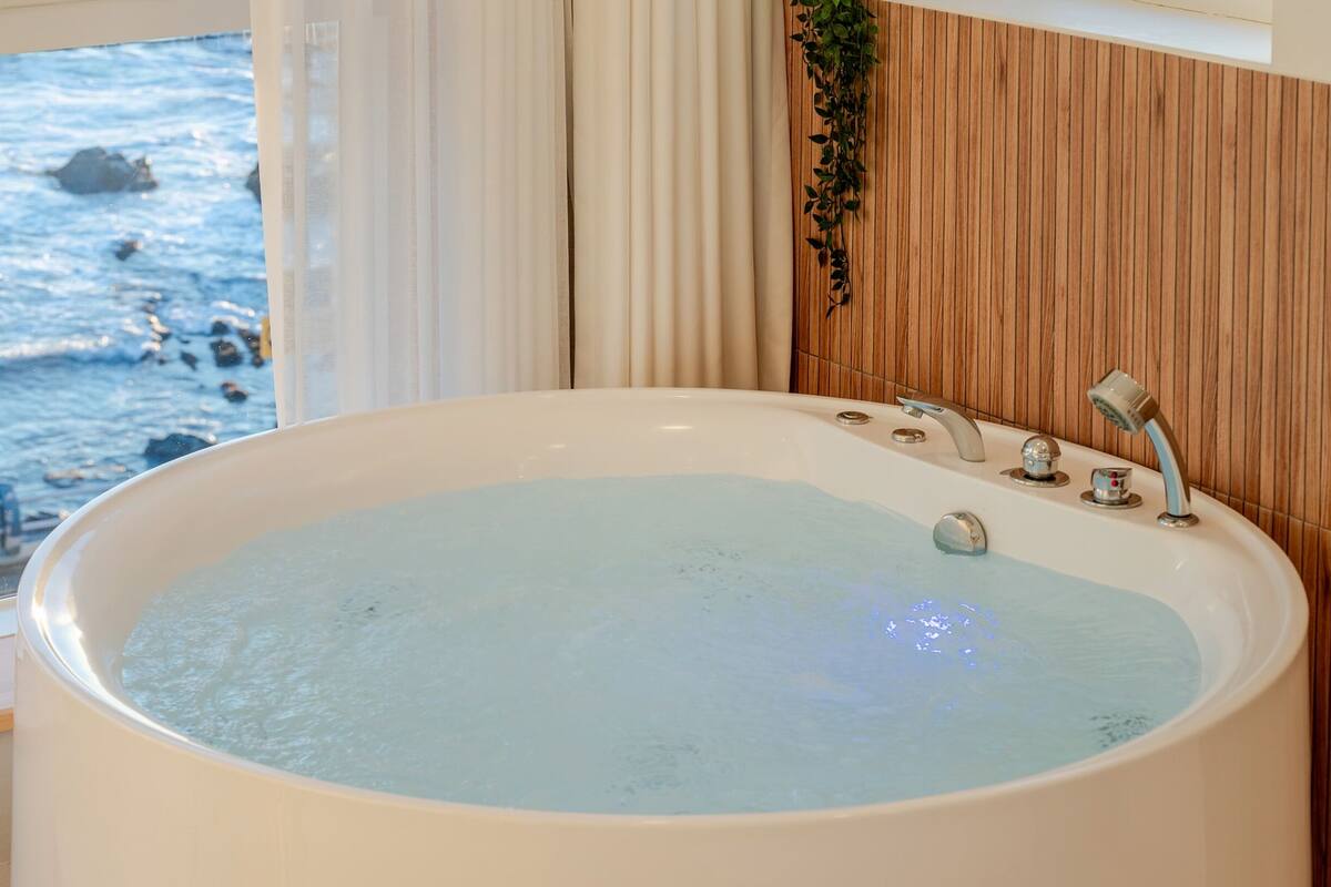 A large bathtub with multiple faucets, a view of the ocean through a window, and a wooden panel wall.