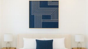 Une chambre à coucher avec un grand lit, deux tables de chevet avec des lampes, un ventilateur de plafond et une œuvre d’art géométrique accrochée au mur.