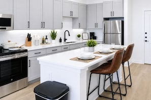 A modern kitchen with white cabinetry, stainless steel appliances, a central island with stools, and a black trash bin. - Qeen Bed · Near Med Center · Long Worker Stays (Sherman)